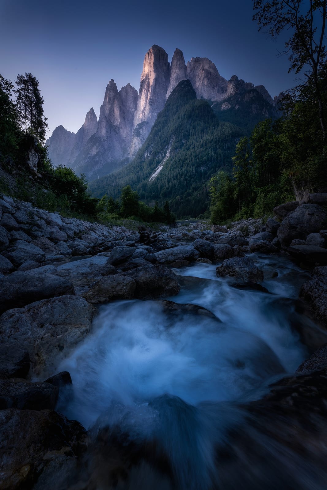 Water swirling in a rock pool below forested slopes and tall rock spires in blue light