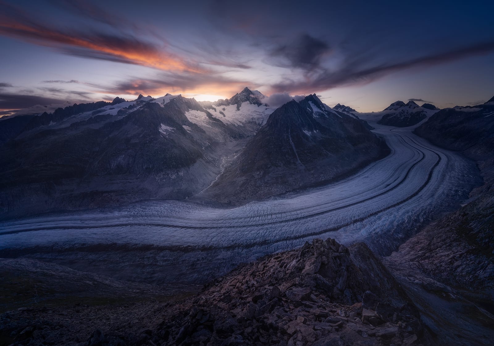 A glacier curving between dark peaks under a streaked sky, the sun low on the horizon