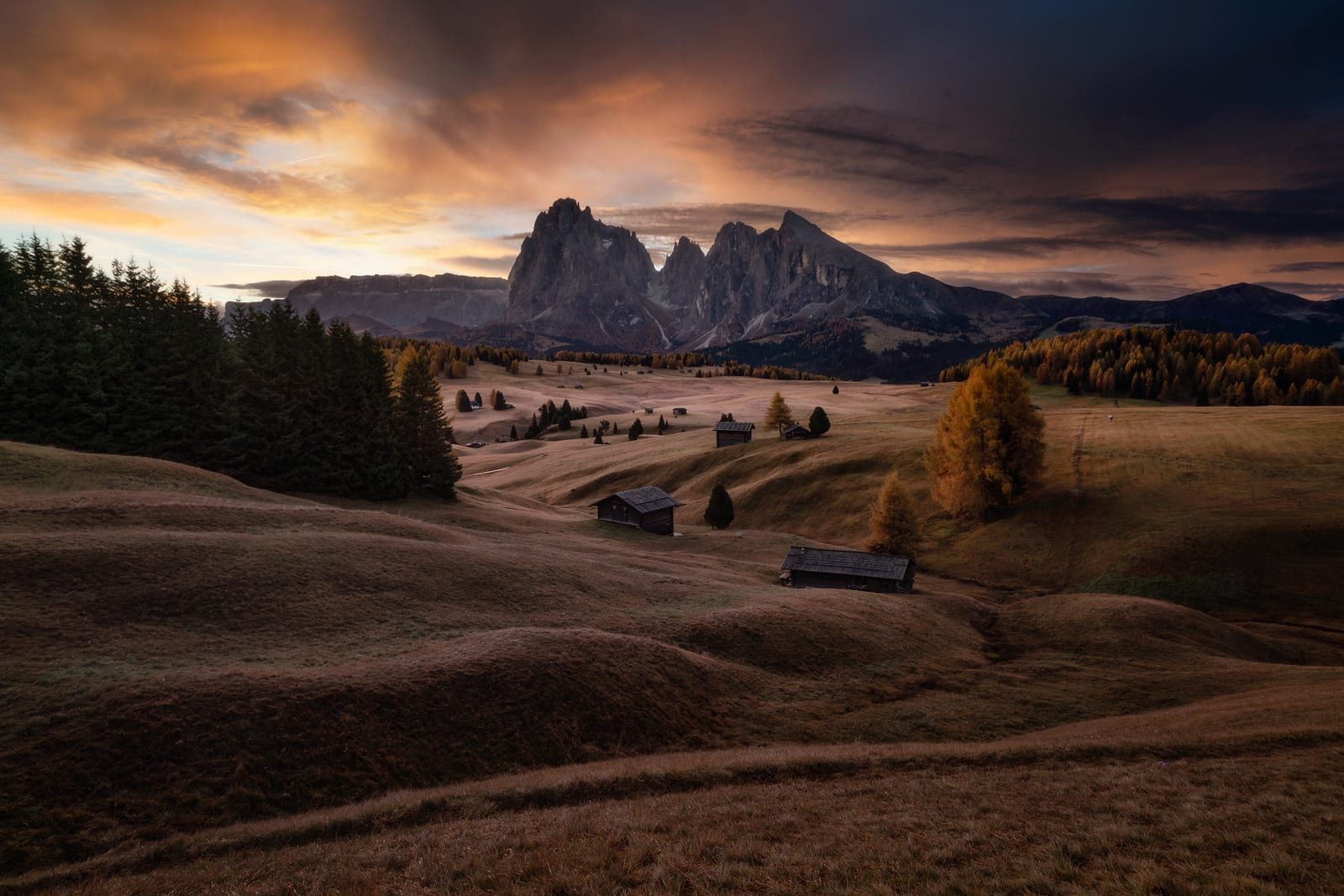 Rolling autumn meadows and wooden huts below jagged peaks under a burning sky