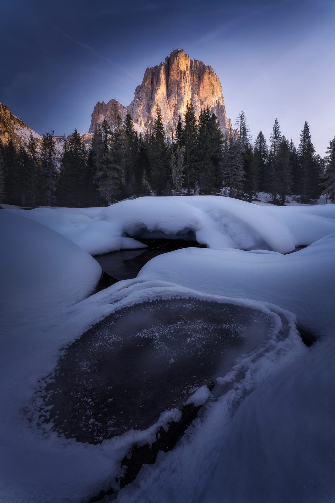 A half-frozen pond in deep snow below pine trees and a rock peak catching the sun