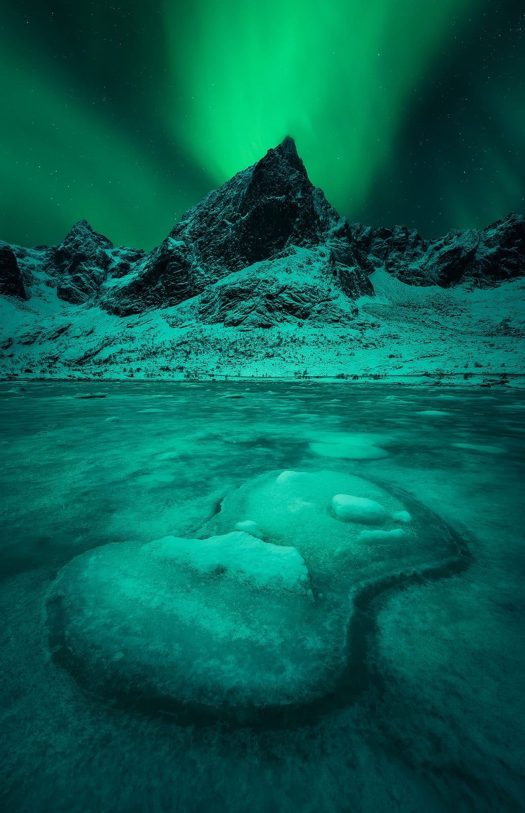 A green aurora above a dark pointed peak, reflected in a frozen lake