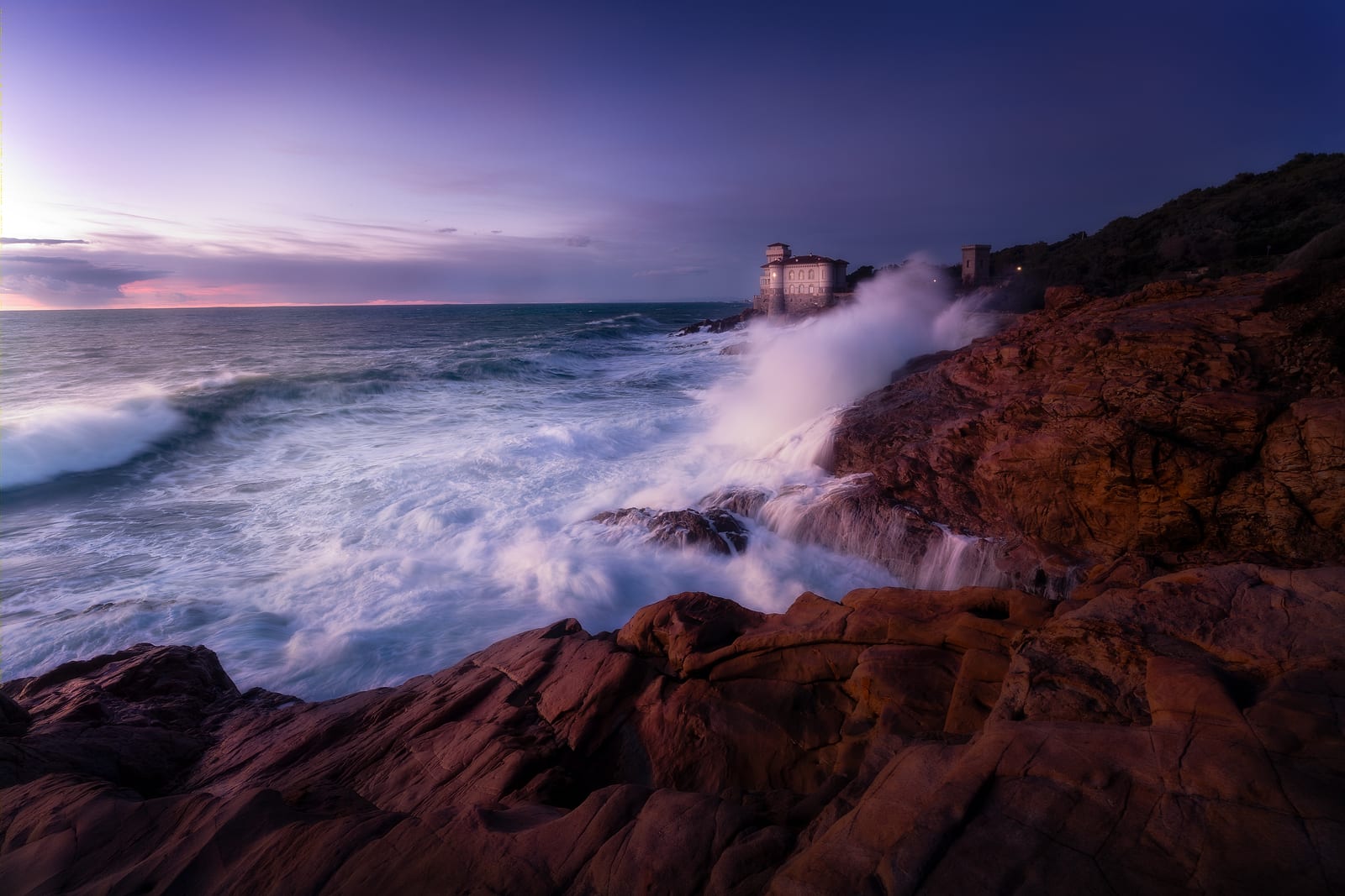 Storm waves breaking on red rocks below a seaside castle in fading light
