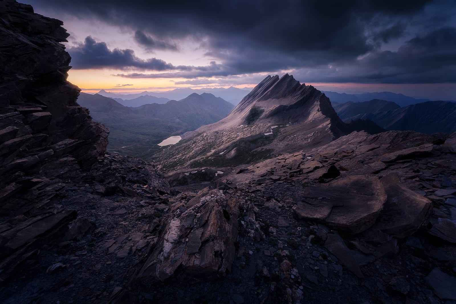 A sharp rock ridge and a small lake below dark clouds, a band of light on the horizon