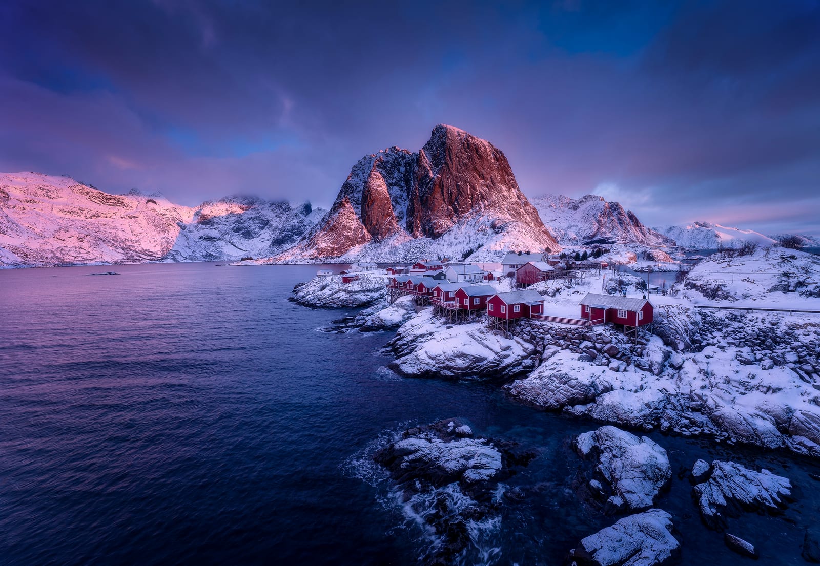 Red fishing cabins on snowy rocks below a peak lit pink