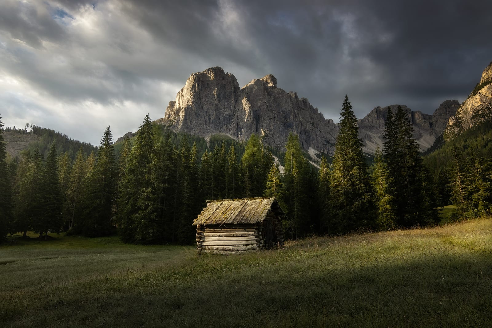 A wooden hut in a meadow below forest and rock peaks under storm clouds