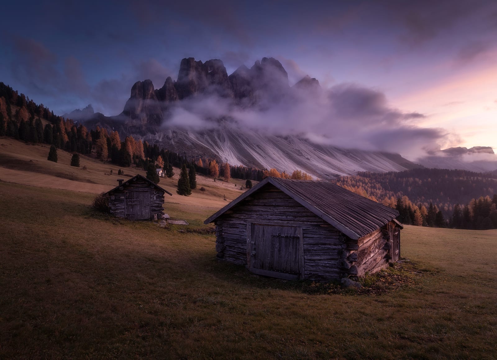 Two log huts in a meadow below rock peaks wrapped in violet cloud