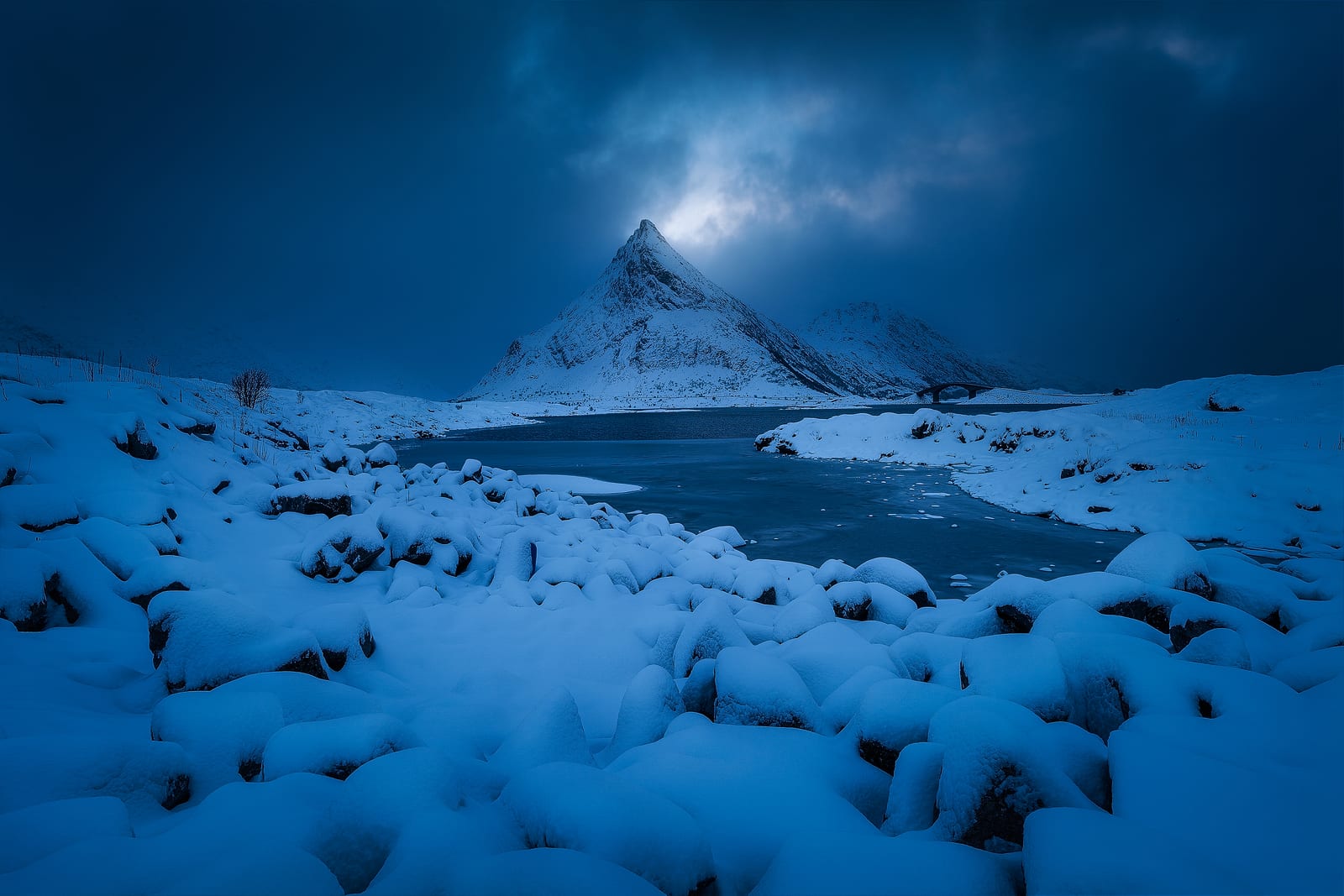 Snow-covered boulders leading to a frozen lake and a pointed peak, a burst of light in a dark sky