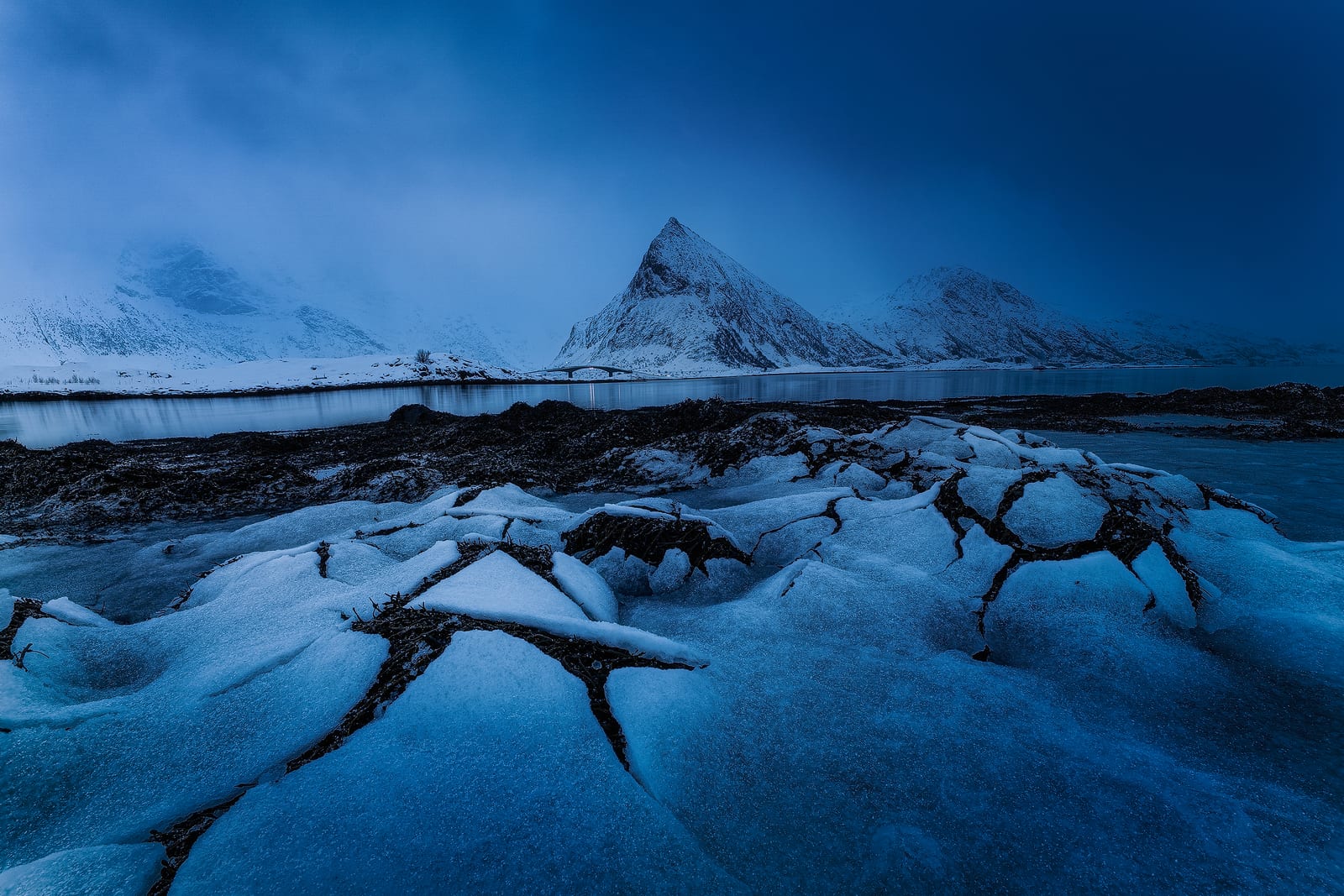 Cracked plates of ice on a shoreline below a pointed snowy peak half lost in fog
