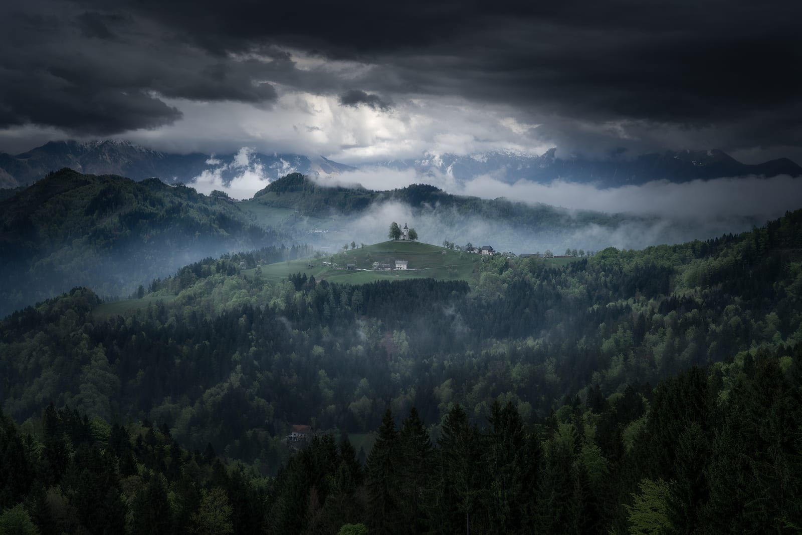 A small church on a green hill lit through dark storm clouds and mist