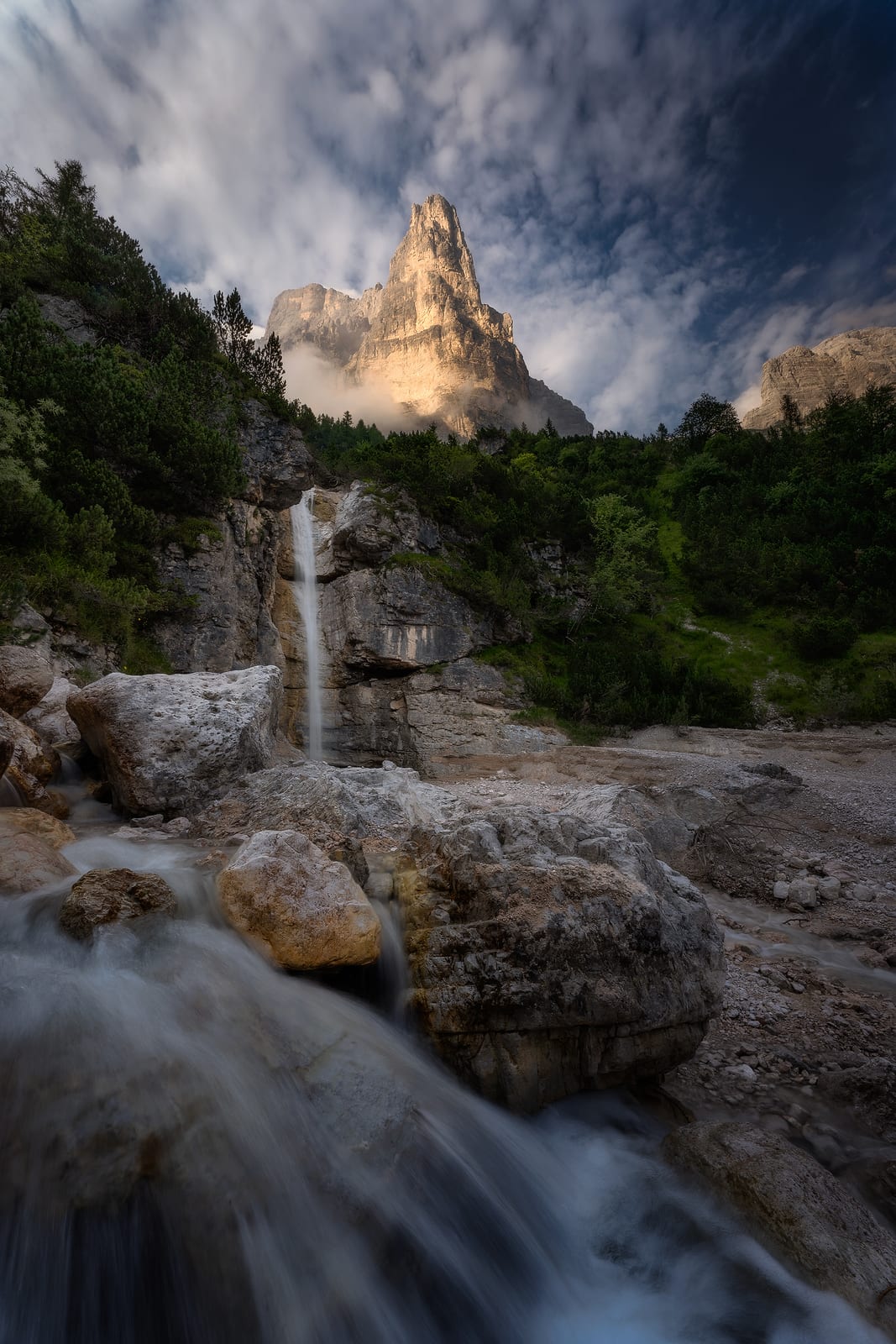 A small waterfall over pale rocks below a tall rock tower and clouds
