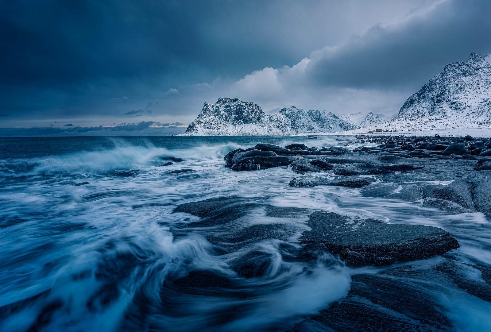 Waves swirling over dark rocks below snowy mountains under storm clouds